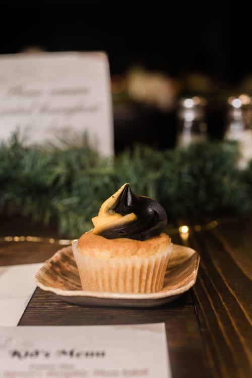 A cupcake with yellow and black frosting on a wooden plate, set against a blurred background of greenery.