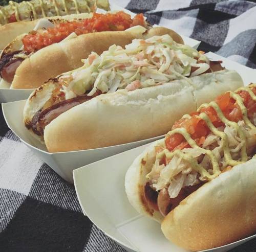 Four hot dogs in buns, topped with various ingredients like coleslaw, tomatoes, and sauces, on a checkered cloth.