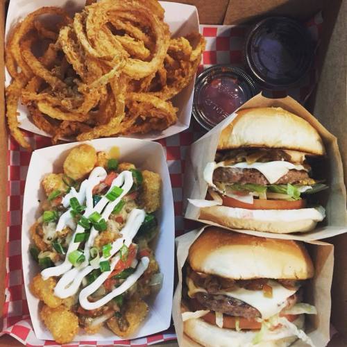 A takeout box filled with onion rings, loaded tater tots, and two burgers with toppings.