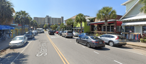 A busy street lined with shops and palm trees, featuring parked cars and a clear blue sky.