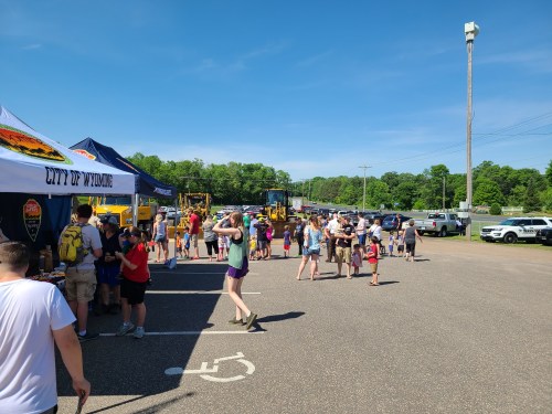 A busy outdoor event with people gathered around tents and vehicles in a parking lot on a sunny day.