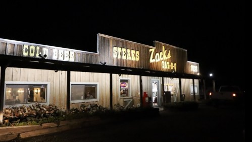 A rustic restaurant at night with a wooden exterior, featuring signs for "Cold Beer" and "Steaks" at Zack's BBQ.