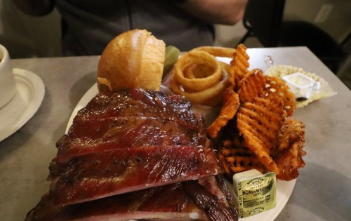 A plate of barbecue ribs with a side of sweet potato fries, onion rings, and a roll, garnished with butter.