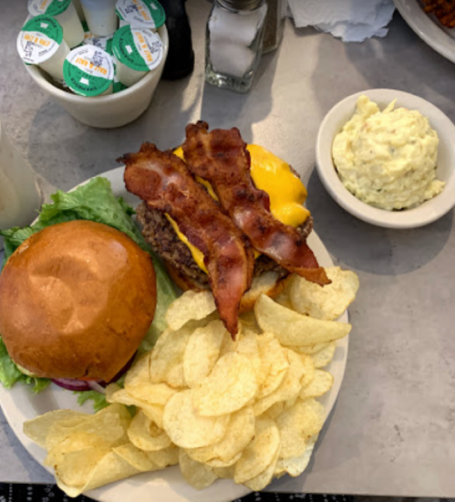 A plate with a cheeseburger topped with bacon, lettuce, chips, and a side of potato salad.