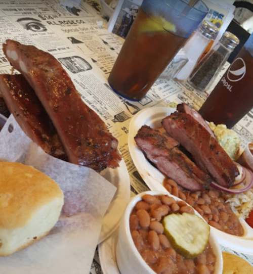A table with BBQ ribs, sliced brisket, cornbread, baked beans, pickles, and iced tea in a casual dining setting.