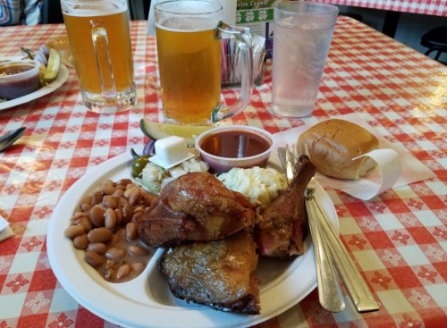 A plate of food with two chicken legs, baked beans, coleslaw, a roll, and two beers on a checkered tablecloth.