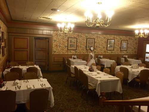 A cozy restaurant interior with floral wallpaper, tables set for dining, and a server attending to the space.