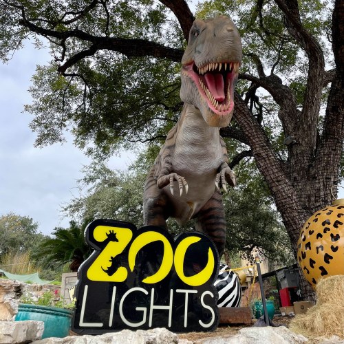 A dinosaur statue next to a sign that reads "Zoo Lights," surrounded by trees and colorful decorations.