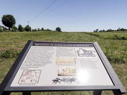 Sign about the beginning of horse racing in Kentucky, with a grassy field and trees in the background under a clear sky.