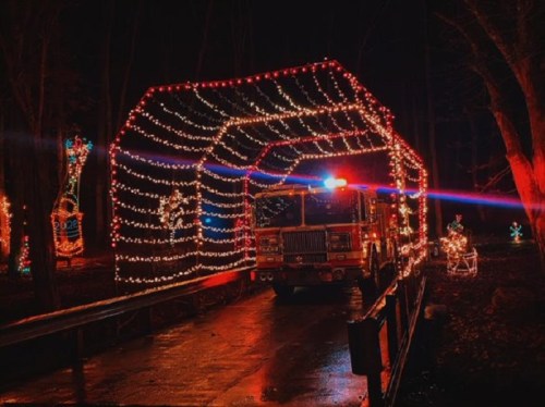 A fire truck decorated with colorful lights drives through a lit archway in a dark, festive forest setting.