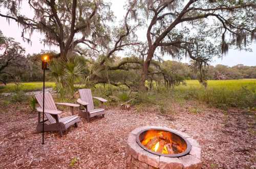 A cozy fire pit surrounded by wooden chairs in a serene outdoor setting with trees and tall grass.