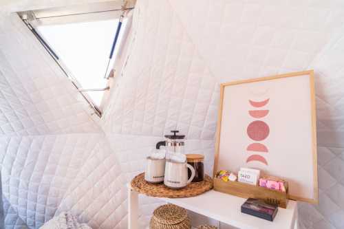 A cozy interior with quilted walls, a decorative print, and a shelf displaying a coffee pot, jars, and snacks.