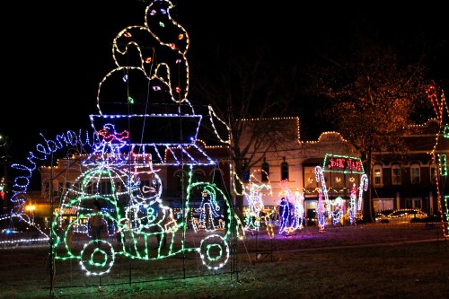 Colorful holiday lights decorate a park, featuring a car with festive characters and a large illuminated bird on top.