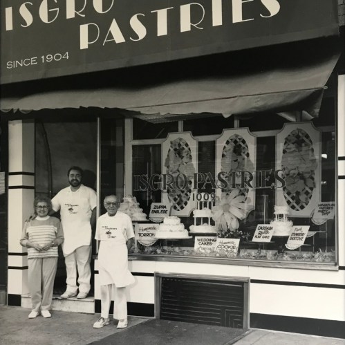 Black and white photo of Isgro Pastries storefront with two people and a man in an apron, showcasing various pastries.