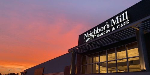 Sign for Neighbor's Mill Bakery & Café illuminated against a colorful sunset sky.