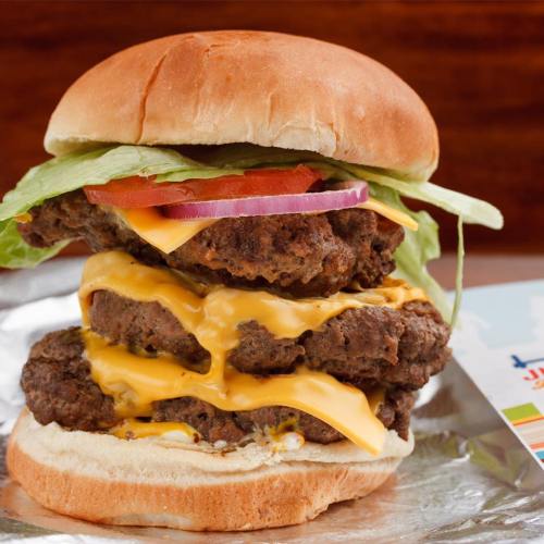 A stacked cheeseburger with three beef patties, melted cheese, lettuce, tomato, and onion in a toasted bun.