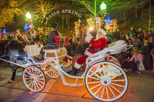 Santa Claus waves from a decorated horse-drawn carriage during a festive parade, surrounded by a cheering crowd.