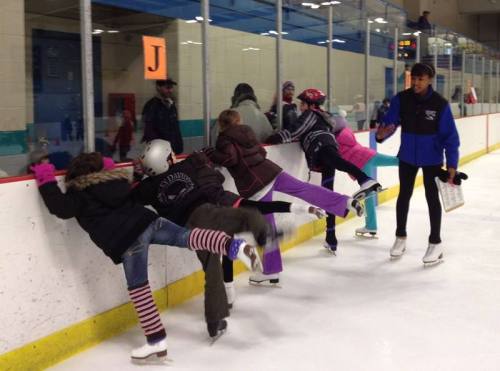 A group of children in winter clothing practice skating at an ice rink, with an instructor nearby.