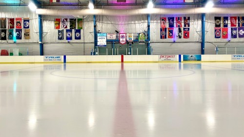 An empty ice rink with colorful banners hanging from the walls and bright overhead lights illuminating the surface.