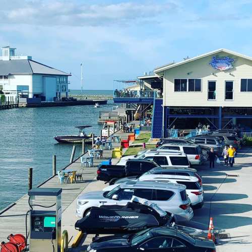 A waterfront scene with parked cars, a marina, and a restaurant building along the shore under a blue sky.