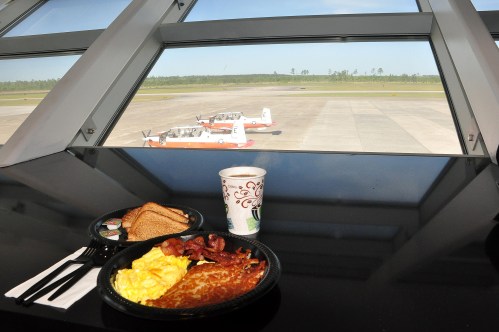 A breakfast plate with eggs, bacon, and toast sits on a table, overlooking small planes outside a large window.
