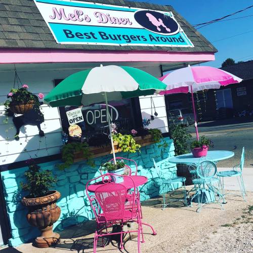 A colorful diner exterior with a sign reading "Mel's Diner," featuring outdoor seating and umbrellas.