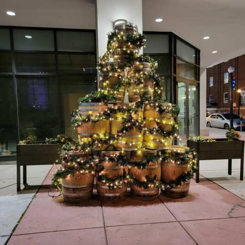 A decorative display of stacked barrels adorned with lights and greenery, set against a modern building backdrop.