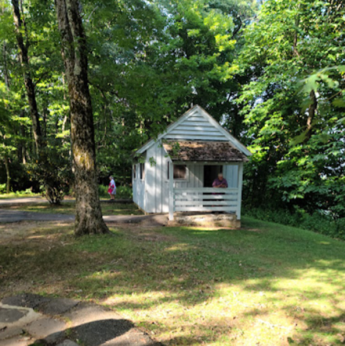A small white cabin surrounded by trees, with a person visible on the porch and another in the background.