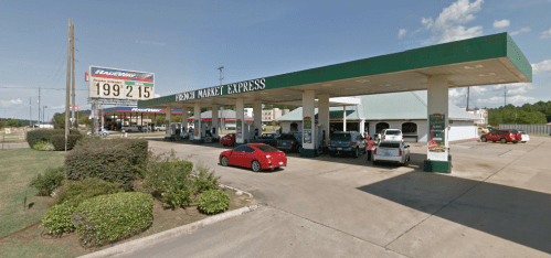 A gas station with multiple pumps, cars parked, and a convenience store under a green canopy on a sunny day.