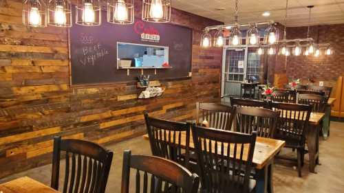 Cozy restaurant interior with wooden walls, tables, and hanging lights, featuring a chalkboard and a kitchen view.