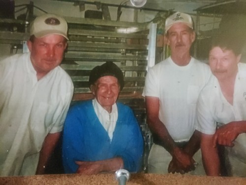 A group of four people, including an elderly woman, posing together in a kitchen setting.