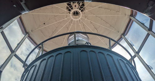 View from inside a lighthouse, looking up at the lantern room and glass dome against a cloudy sky.