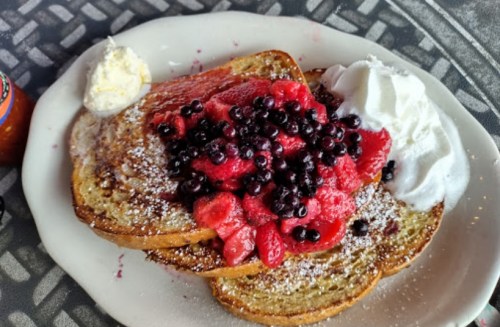 A plate of French toast topped with mixed berries, whipped cream, and a dollop of butter, dusted with powdered sugar.