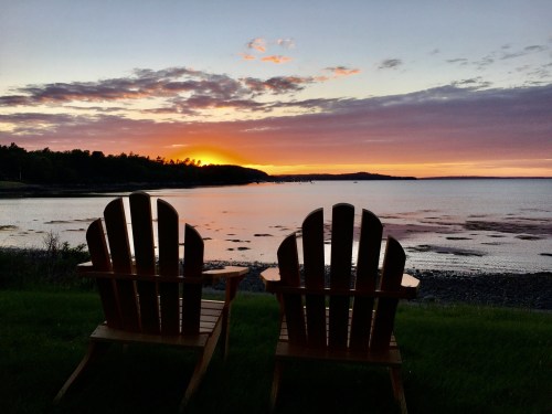 Two wooden Adirondack chairs facing a serene sunset over a calm bay, with colorful clouds reflecting on the water.