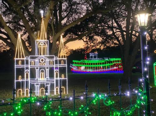 Colorful holiday lights depict a church and a boat against a sunset backdrop, surrounded by trees.