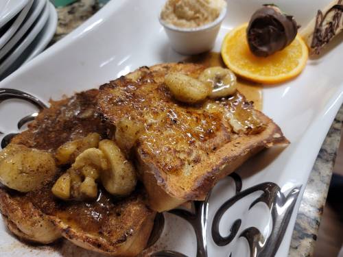 A plate of French toast topped with caramelized bananas, syrup, and a side of orange and chocolate-dipped treat.