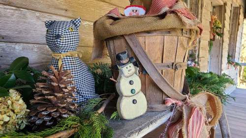 A rustic display featuring a checkered cat, a wooden snowman, a basket, pinecones, and greenery on a wooden surface.