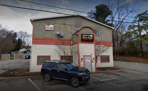 A two-story building with a red and gray exterior, featuring a sign for "Ala Rota" and a parked black SUV in front.