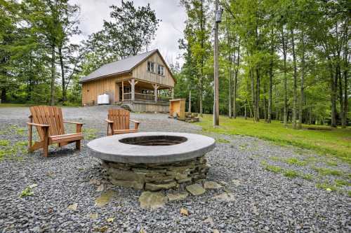 A cozy cabin surrounded by trees, featuring a stone fire pit and two wooden chairs on a gravel area.