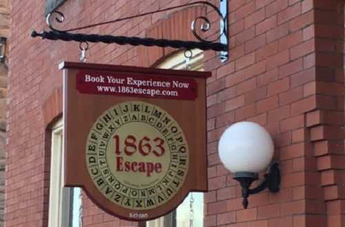 Sign for "1863 Escape" with a circular design featuring letters, hanging from a brick wall near a light fixture.