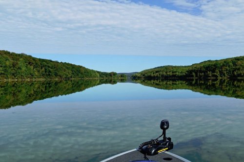 A serene lake surrounded by lush greenery, reflecting the sky and trees, viewed from a boat.