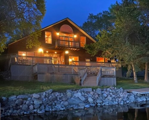 A cozy wooden cabin by a lake, illuminated at night, surrounded by trees and a stone shoreline.
