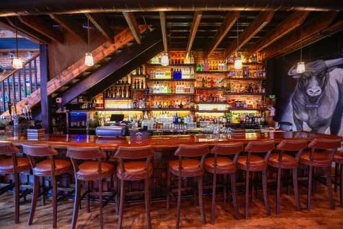 A rustic bar with wooden beams, shelves filled with colorful bottles, and leather stools arranged along the counter.