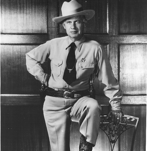 A man in a sheriff's uniform poses confidently with one leg on a chair, wearing a wide-brimmed hat and a badge.