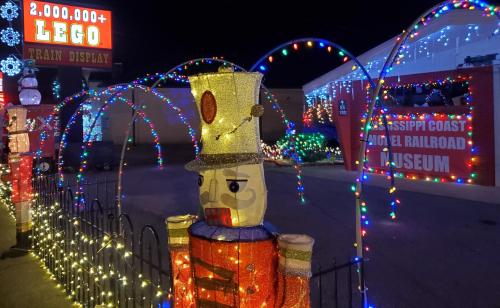 Colorful holiday lights illuminate a LEGO train display with a toy soldier figure in the foreground.