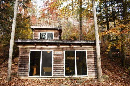 A rustic wooden cabin surrounded by autumn foliage and trees, featuring large glass doors and a two-story design.