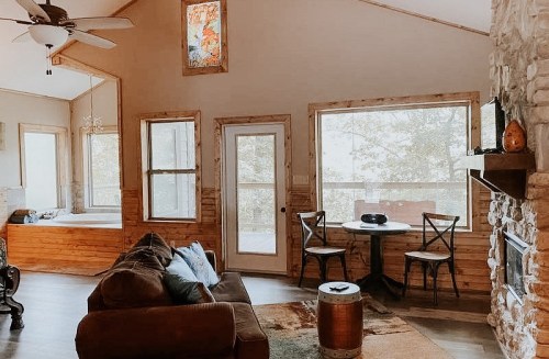 Cozy living room with wooden accents, large windows, a stone fireplace, and a small dining table.