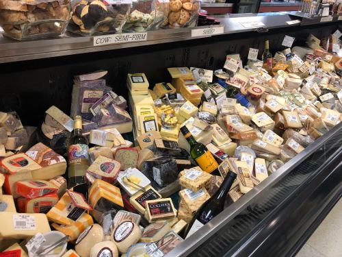 A variety of cheeses displayed on a market shelf, with bottles of wine among the assortment.