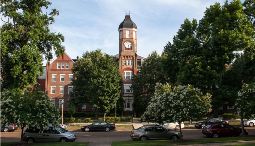 Historic building with a clock tower surrounded by trees and parked cars on a sunny day.