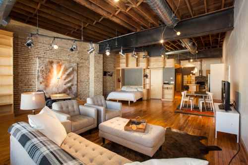 A modern loft with exposed brick walls, wooden floors, a living area, kitchen, and a bed in an open layout.